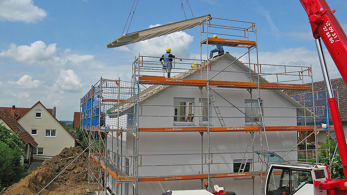 Auf ein eingerüstetes Wohnhaus wird ein Dachelement von einem Kran herabgelassen.