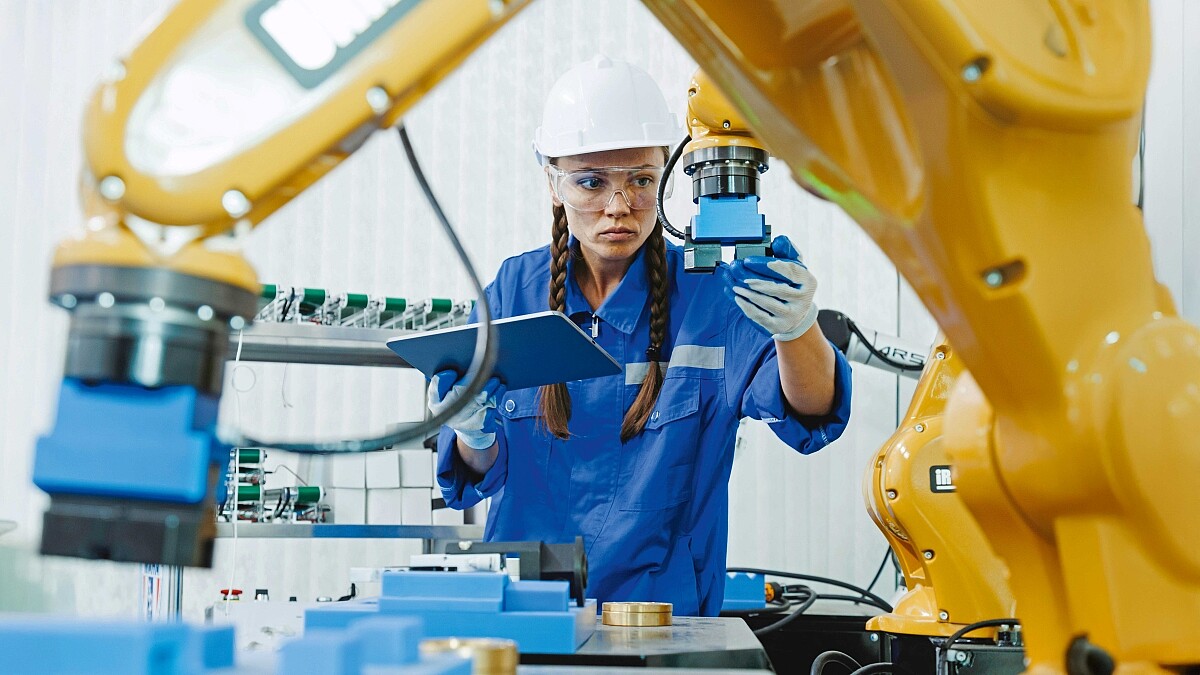 Eine Frau mit Schutzhelm und Arbeitshandschuhen hält ein Tablet in der Hand und steuert eine Maschine.