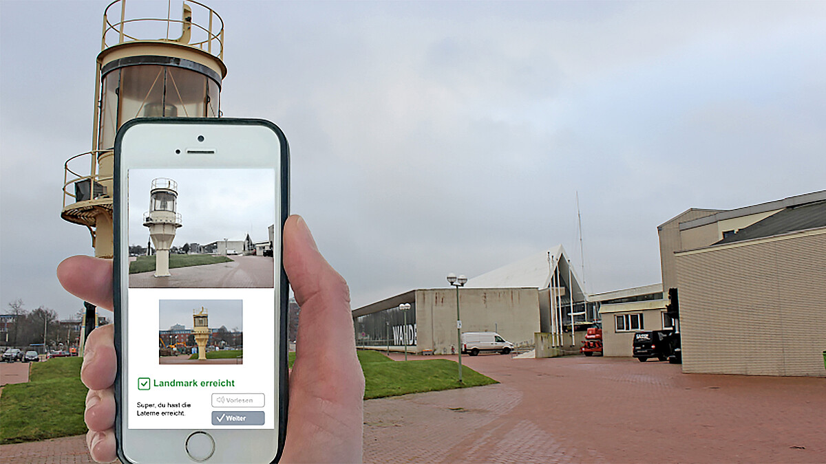 Vor einem niedrigen Leuchtturm hält eine Hand ein Mobiltelefon in die Höhe. Auf dem Handydisplay ist der Leuchtturm zu sehen und die Information „Landmark erreicht“.