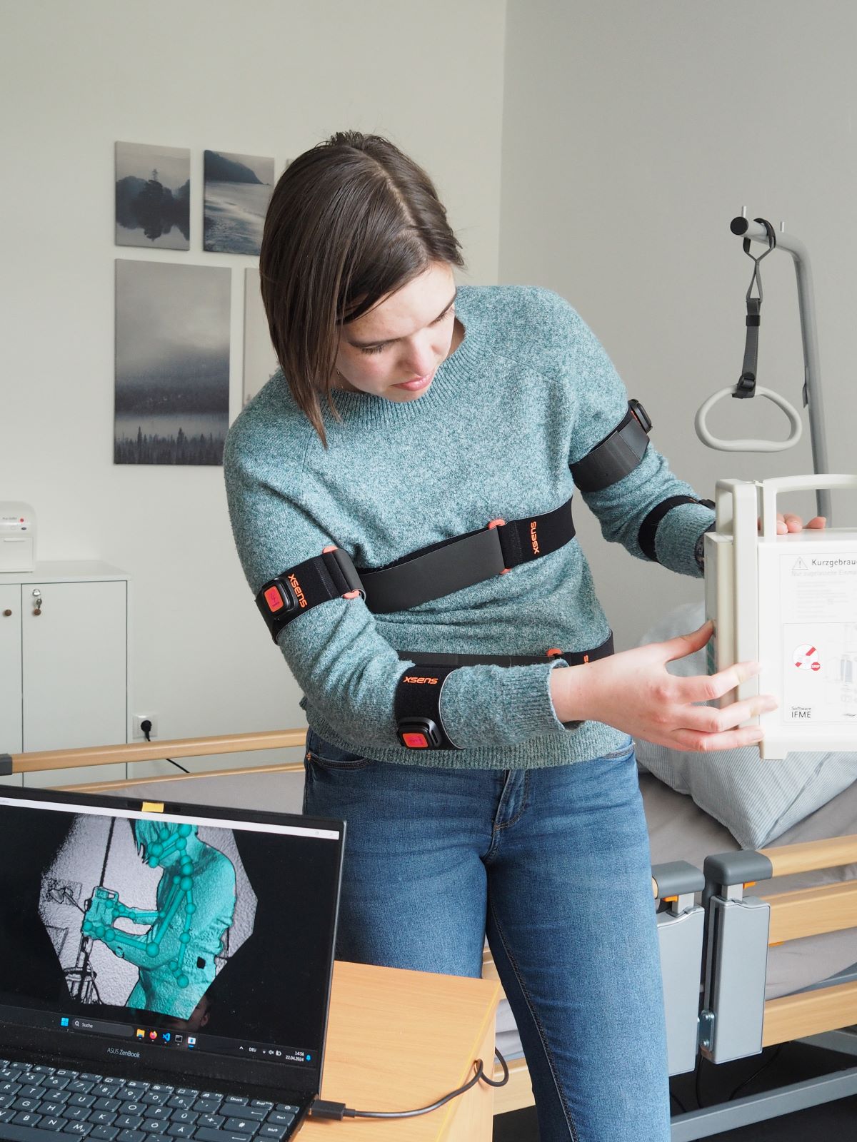 Eine Person mit Sensorbändern um Arme und Oberkörper überprüft eine Apparatur im Krankenzimmer. Auf einem Laptop wird die gefilmte Situation vermessen.